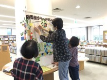 奥に本棚がたくさんある部屋で「図書館からのお知らせ7月」と書かれたタペストリーを二人の女性が作成しており、一人がそれを見守っている写真