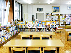 大きな窓からの光や照明に照らされて明るい室内を囲むように沢山の本棚が置かれ、木目調の長机と椅子が並んでいる図書館の写真