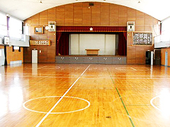 バスケットボールコートや講壇があり、木目調の床の、広く大きな体育館内の写真