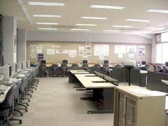 沢山のパソコンが置かれた机が部屋を囲むように並び、白い壁で沢山の電灯に照らされている明るいパソコン教室の写真
