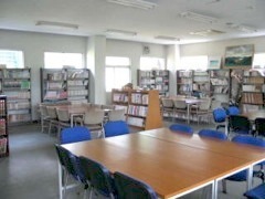 茶色い四角状のテーブルと青い椅子が置かれ、沢山の本棚に囲まれた図書室の写真