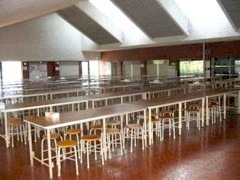 沢山の長机と椅子が立ち並び、三角状の屋根から光が入ってきている食堂の写真