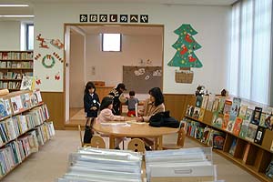 木製の丸い机を囲んで読み聞かせをしている女性とそれを聞いている子ども達の様子の写真