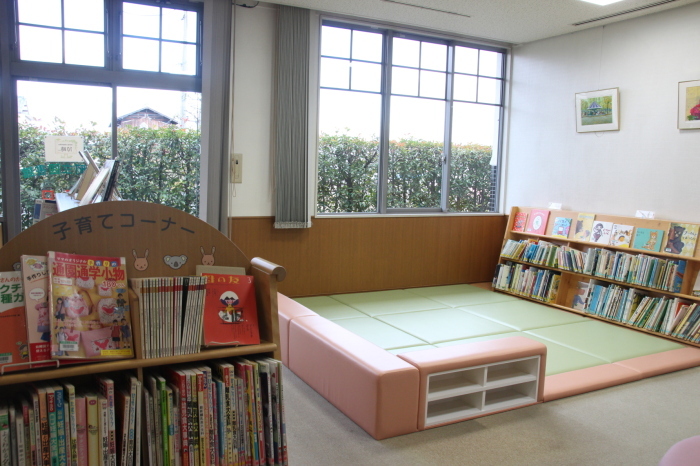 茶色のソファーで囲まれ、靴を脱いでくつろげる緑色の床が正方形状に広がっているスペースと「子育てコーナー」と書かれた本棚に本が並んでいる写真