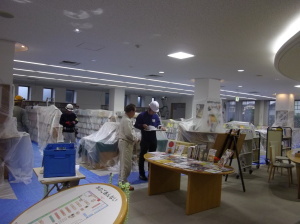 書架にビニールが掛けられ床に青い養生シート敷かれている図書館で、作業服を着た2人が打ち合わせをしている写真