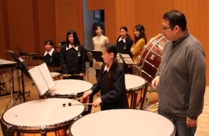 大人の男性が立っている横でたくさんのドラムを叩く女の子の学生の写真