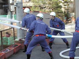 3人のヘルメットを被った消防団員が2本のホースを持って協力して放水している写真