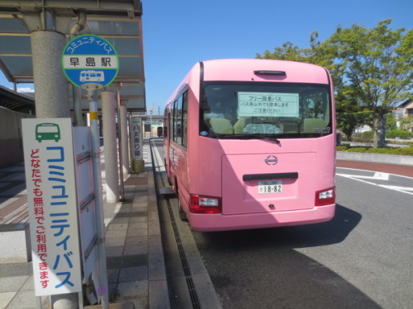 「コミュニティバス どなたでも無料でご利用できます」と書かれた看板が置いてある、屋根のある早島駅停留場でピンクのバスが停車している写真