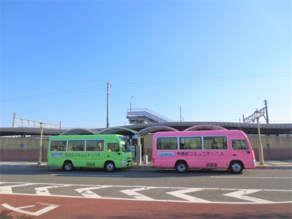 青空の下、黄緑色とピンク色のバスが縦並びに並んで停留所に停車している写真