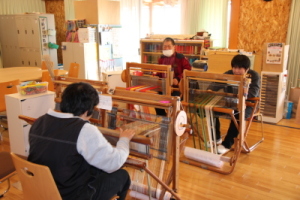 木目調の床で日が差し込む明るい部屋に、椅子や机が立ち並び、その椅子に3人が座っている写真