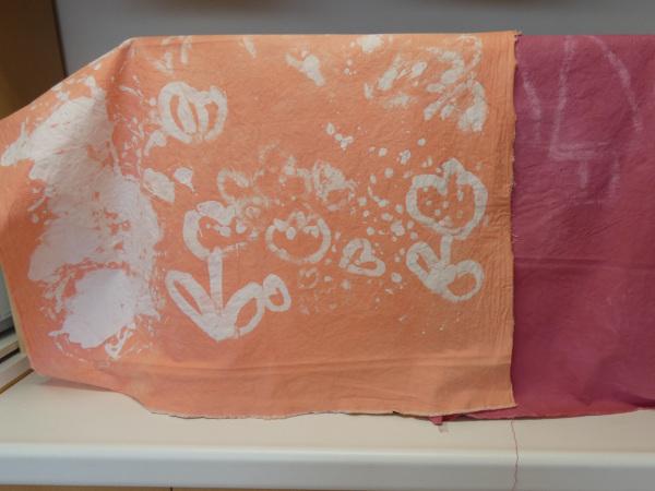 ろうけつ染めで作られたオレンジ色の布に花の絵が描かれている作品の写真
